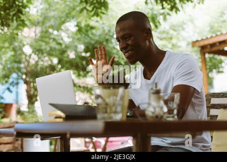 Smiling and glad multiracial man with laptop, raising arm, greet someone, looking at monitor. E-learning webinar meeting Stock Photo