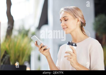 Content blond young woman in casual sweater drinking coffee from takeout cup and texting message on smartphone Stock Photo