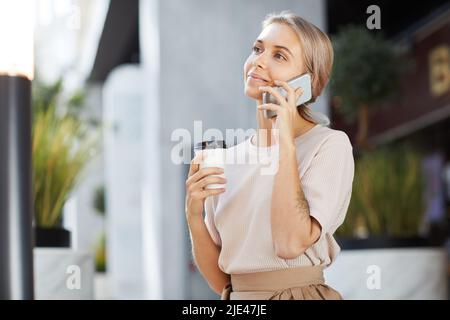 Smiling pretty young woman with tattoo on arm standing in lobby and communicating on phone while drinking coffee Stock Photo