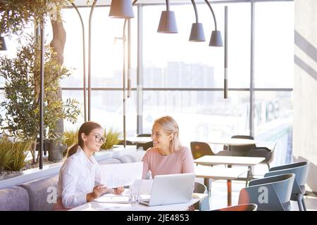 Positive attractive young businesswomen sitting at table in cozy restaurant and analyzing graphs while making sales forecast Stock Photo