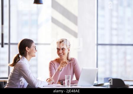 Cheerful beautiful marketing specialists in formalwear sitting at table in office and using laptop while creating advertising plan for new product Stock Photo