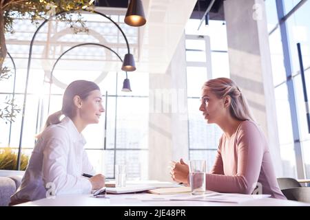 Serious confident young businesswomen sitting at table with papers and talking to each other while having lunch together Stock Photo