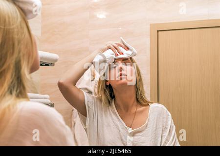 Headache, migraine. Portrait reflection in mirror of young girl making cold compress with towel on her head. Woman standing in front of mirror in
