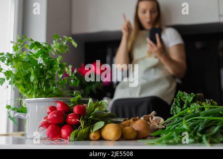 Fresh vegetable on table. Plus size blond woman in apron using smartphone in the kitchen. Find diet recipe in mobile app Stock Photo