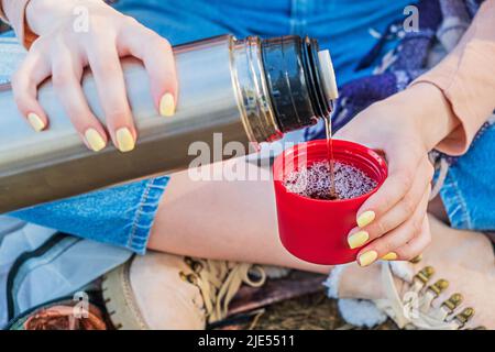 Cropped young girl sitting in a tent in the woods pours hot tea into a thermos cup. Mental health. Time alone, contemplation. Fall or spring time Stock Photo