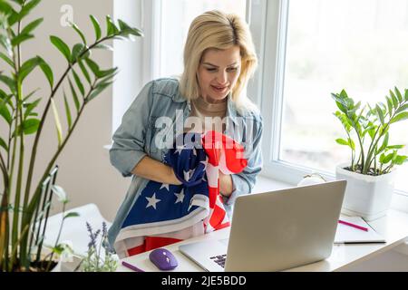A woman sport fan covered with american flag sits on sofa and watches sport match on laptop in living room and cheers for the american team Stock Photo