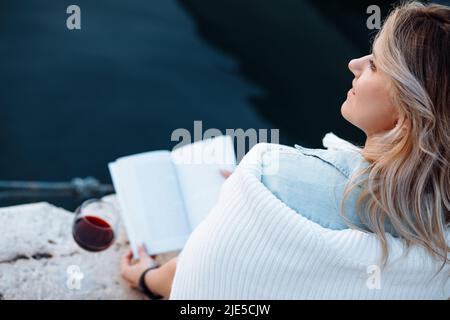 beautiful young woman is sitting on pier with glass of red wine and book having great time on vacation , happy evening. Stock Photo