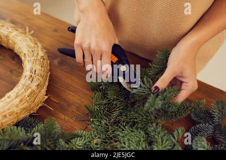 Close-up of unrecognizable female florist standing at wooden desk and cutting coniferous branch while making Christmas wreath Stock Photo