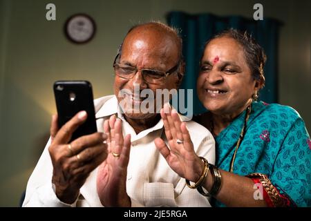 happy senior couple busy on making video call using smartphone at home - concept of technology, bonding and connection Stock Photo