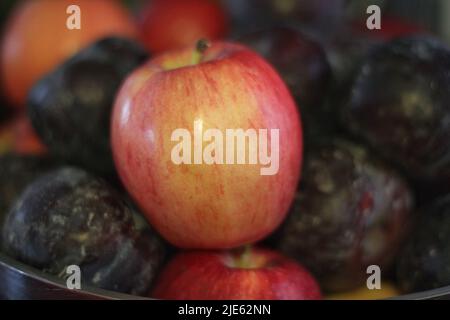 Delicious red apple sitting among a bunch of tasty purple plums in a fruit bowl. Stock Photo
