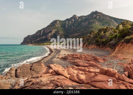 The grey pebble beach Coccorocci framed by red porphyry rocks and the woods around Monte Cartucceddu on the east coast of Sardinia, Ogliastra, Italy Stock Photo