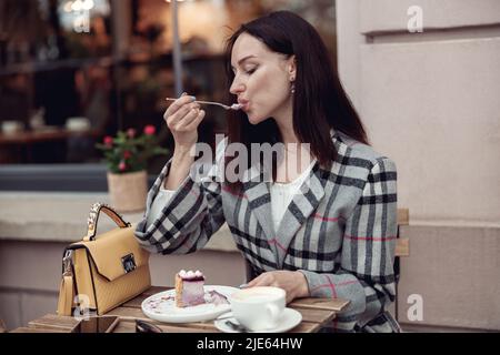 Beautiful brunette female enjoying cheesecake pie with coffee outdoor at cafe. Rest in city. Stock Photo