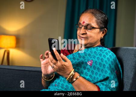 Senior woman busy using mobile phone while sitting on sofa at home - concepts of technology, relaxation and social media Stock Photo