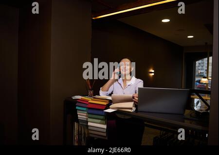 Pleasant female sales representative talking on mobile phone while standing at desk with laptop and upholstery fabric samples, taking orders in furnit Stock Photo