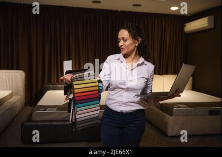 Creative woman professional decorator working on new home design project, holding laptop in her hands and choosing upholstery fabrics, standing agains Stock Photo