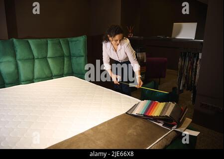 Interior designer using measuring tape measures the length of the mattress and the soft bed to order new upholstery. Upholstery fabric samples lie on Stock Photo