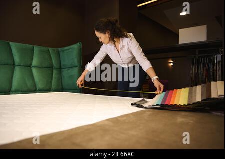 Beautiful Hispanic woman - saleswoman holding a measure tape measuring the orthopedic mattress dimensions in the furniture design showroom. Upholstery Stock Photo