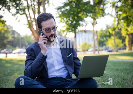 Young entrepreneur sitting in the park continuing to work using cell phone and PC. Stock Photo