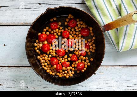 fried cherry tomatoes with chickpeas cooked in a cast iron pan on a light wooden background, top view Stock Photo