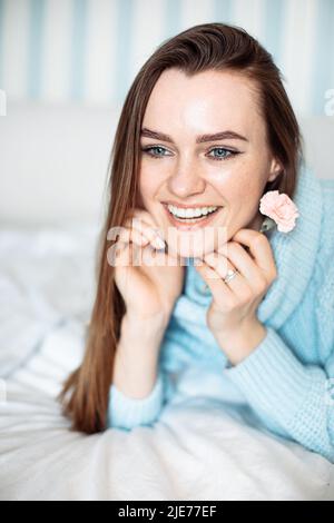 Smiling happy woman lying on bed in cozy sweater, holding delicate flower in hand, closeup portrait  Stock Photo