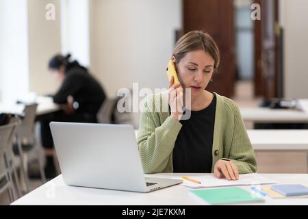 Middle-aged woman busy with documents talking on cellphone working in campus open space. Education.  Stock Photo