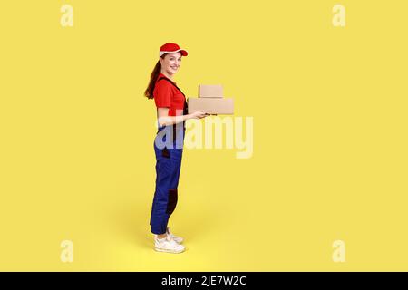 Side view portrait of courier woman standing with two parcels in hands, looking at camera, delivery service, wearing overalls and red cap. Indoor studio shot isolated on yellow background. Stock Photo