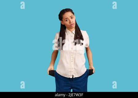 Unemployment and bankruptcy. Upset poor woman with dreadlocks turning out empty pockets, worried about debts, no cash for living, wearing white shirt. Indoor studio shot isolated on blue background. Stock Photo