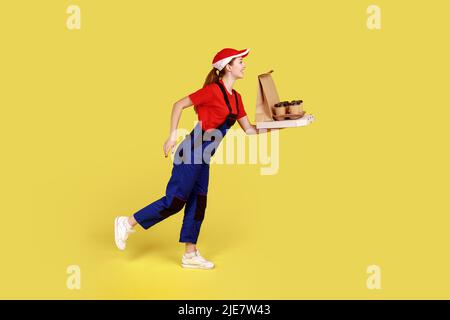 Side view portrait of delivery woman being hurry to deliver coffee with pizza, fast delivery service, wearing overalls and red cap. Indoor studio shot isolated on yellow background. Stock Photo
