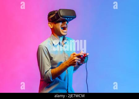 Side view of excited man in shirt in VR glasses playing video game, using simulator for education, yelling emotionally. Indoor studio shot isolated on colorful neon light background. Stock Photo