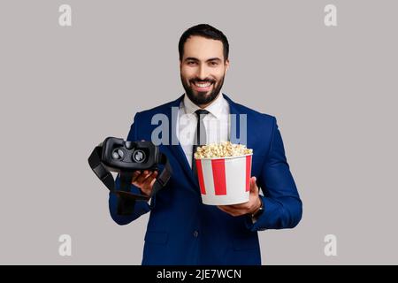 Positive bearded man holding VR headset in hands, using augmented reality eyeglasses being in virtual reality, watching movie with popcorn. Indoor studio shot isolated on gray background. Stock Photo