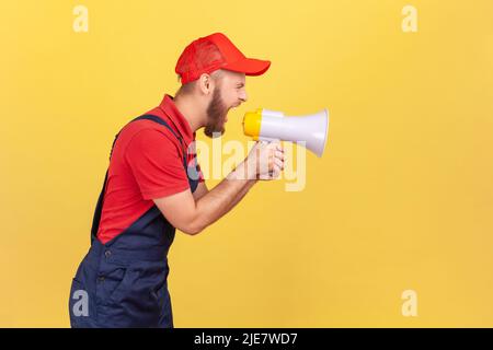 Side view portrait of angry bearded worker in blue uniform and red cap, holding megaphone and screaming with aggressive expression, protesting. Indoor studio shot isolated on yellow background. Stock Photo