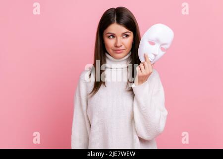 Portrait of young beautiful adult female with dark hair looking away, holding in hands white mask, wearing white casual style sweater. Indoor studio shot isolated on pink background. Stock Photo