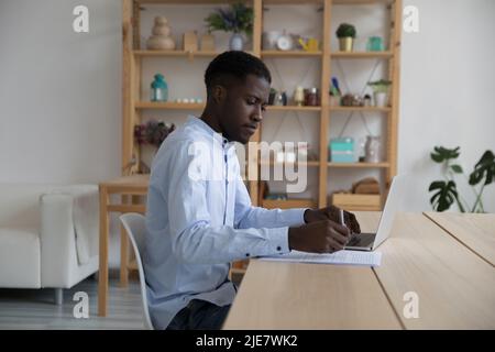 African businessman writes notes, working seated at desk with laptop Stock Photo