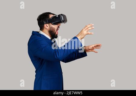 Side view of man in vr headset, playing virtual reality game with shocked facial expression, outstretching hands, wearing official style suit. Indoor studio shot isolated on gray background. Stock Photo