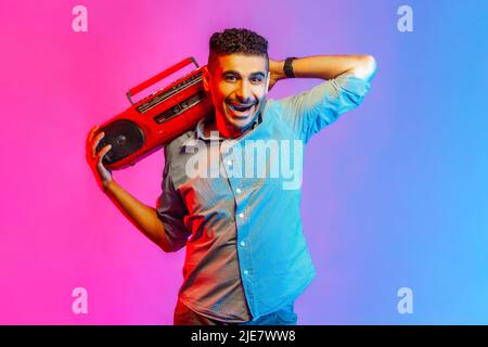 Portrait of man in shirt listening to music holding red tape recorder on shoulder, looking at camera with toothy smile. Indoor studio shot isolated on colorful neon light background. Stock Photo