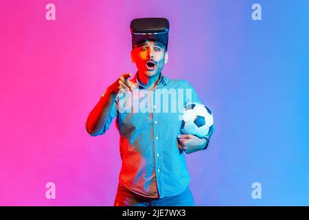 Portrait of astonished surprised man in shirt wearing virtual reality glasses, posing with soccer ball, pointing finger to camera. Indoor studio shot isolated on colorful neon light background. Stock Photo