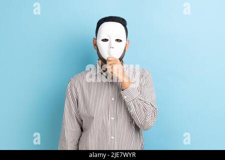 Portrait of anonymous unknown businessman covering her face with white mask, hiding her real personality, anonymity, wearing striped shirt. Indoor studio shot isolated on blue background. Stock Photo
