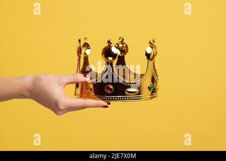 Closeup of woman hand holding golden crown, concept of awards ceremony, privileged status, superior position. Indoor studio shot isolated on yellow background. Stock Photo