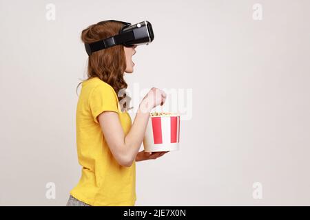 Young surprised shocked girl in yellow T-shirt using augmented reality eyeglasses being in virtual reality, watching horror movie and eating popcorn. Indoor studio shot isolated on gray background. Stock Photo