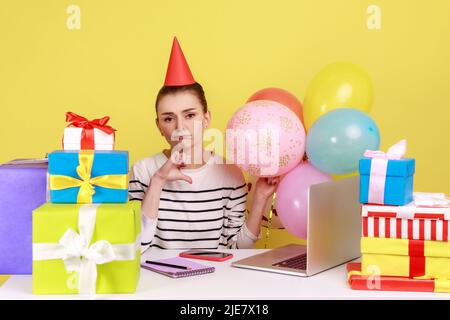 Sad woman in party cone celebrating birthday alone at workplace among many presents and air balloons, showing thumb down, dislike party. Indoor studio studio shot isolated on yellow background. Stock Photo