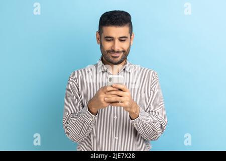 Portrait of cheerful businessman using cellphone and smiling, reading good news message, enjoying mobile application, wearing striped shirt. Indoor studio shot isolated on blue background. Stock Photo