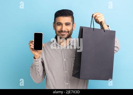 Smiling businessman with beard holding white display smartphone and black paper bag, online shopping, fast delivery, marketplace, wearing striped shirt. Indoor studio shot isolated on blue background. Stock Photo