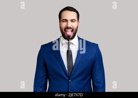 Portrait of playful bearded businessman showing tongue, having fun, fooling around, childish manners, wearing official style suit. Indoor studio shot isolated on gray background. Stock Photo