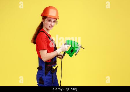 Side view portrait of builder woman working with fretsaw, looking at camera with serious confident expression, wearing overalls and protective helmet. Indoor studio shot isolated on yellow background. Stock Photo