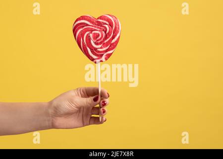 Closeup of woman hand holding appetizing heart shaped candy on stick, big lollipop in arm, confectionery advertising, glucose sweet foods. Indoor studio shot isolated on yellow background. Stock Photo