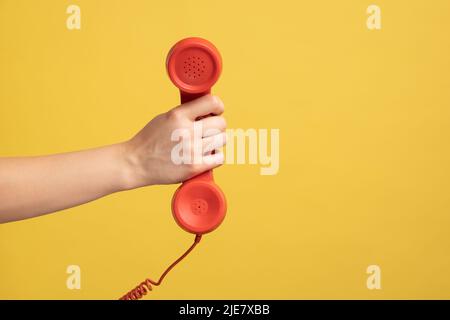 Profile side view closeup of woman hand holding and showing red call telephone handset receiver, call center. Indoor studio shot isolated on yellow background. Stock Photo