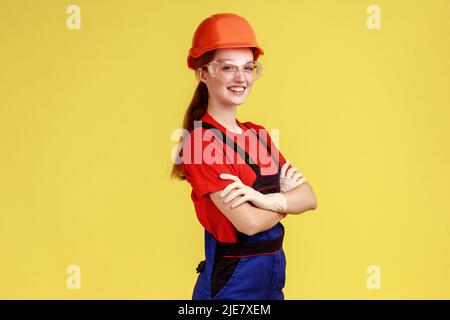 Side view of positive worker woman standing with crossed arms and looking at camera with confident expression, wearing overalls and helmet. Indoor studio shot isolated on yellow background. Stock Photo