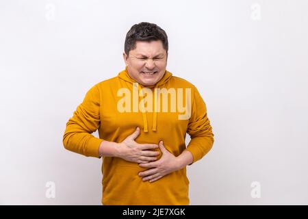 Stomachache. Portrait of sick middle aged man grimacing and suffering from pain in belly, severe abdominal distress, wearing urban style hoodie. Indoor studio shot isolated on white background. Stock Photo