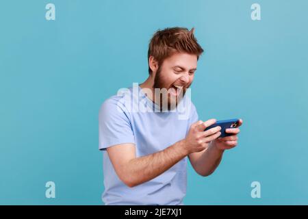 Portrait of handsome young adult bearded man standing, using smartphone and playing mobile game with excited positive face, happy to win. Indoor studio shot isolated on blue background. Stock Photo