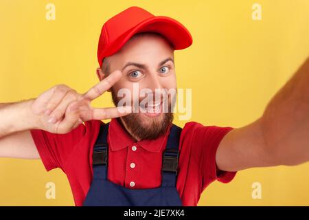 Smiling positive man courier or repairman broadcasting livestream or making point of view photo, showing v sign, wearing blue overalls and red cap. Indoor studio shot isolated on yellow background. Stock Photo